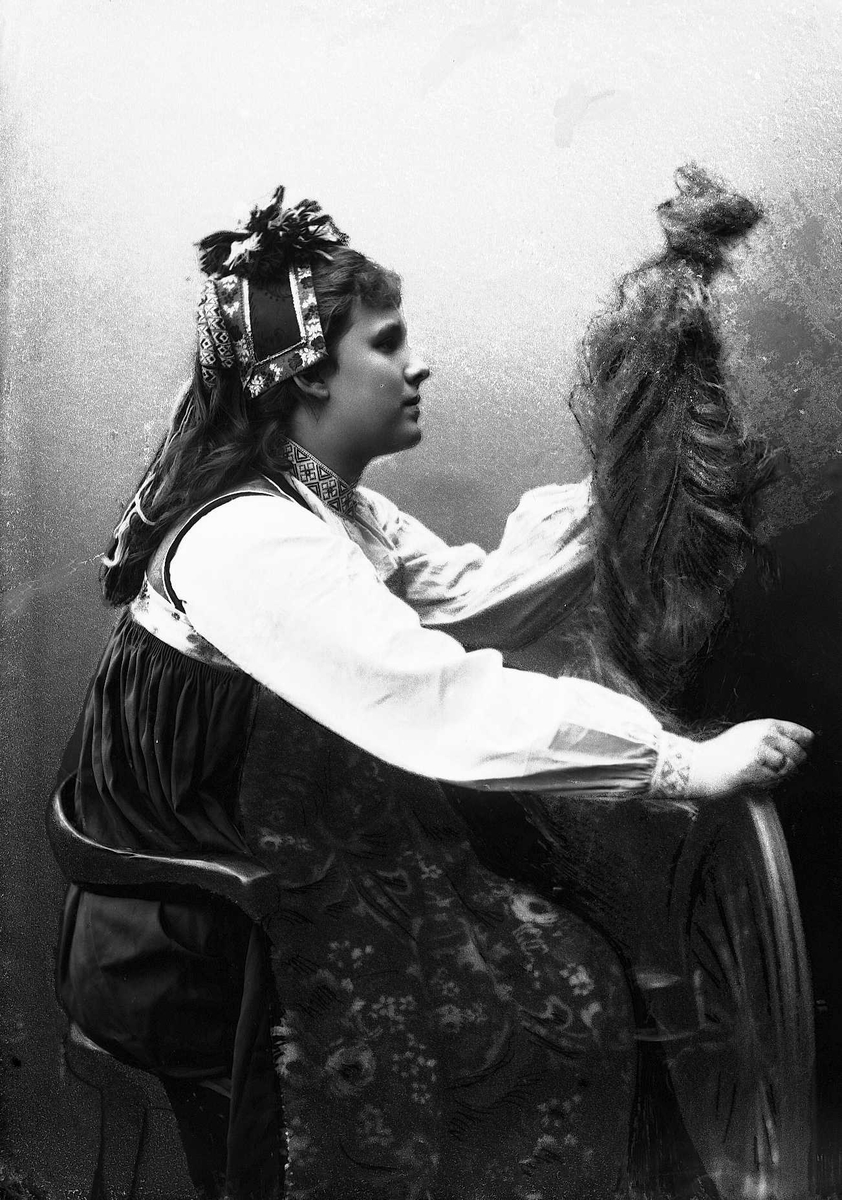 Solveig Lund, Studioportrett av kvinne i drakt og hodeplagg, som sitter ved en rokk. Bakgrunnen er nøytral.
