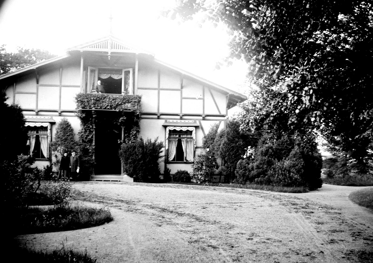 Juliane Fredrikke Langberg, Fam. Hudtwalckers hjem i Blankenese, Hamburg, muligens med hr. og fru Hudtwalcker stående utenfor t.v. og Heinrich C. Hudtwalcker på balkongen over. Frodig hage omkring huset og gårdsplassen. Fam. er svigerfamilie til fotografens niese, Sigrid f. Holm. Fra malerinne Juliane Fredrikke Langbergs (185…
