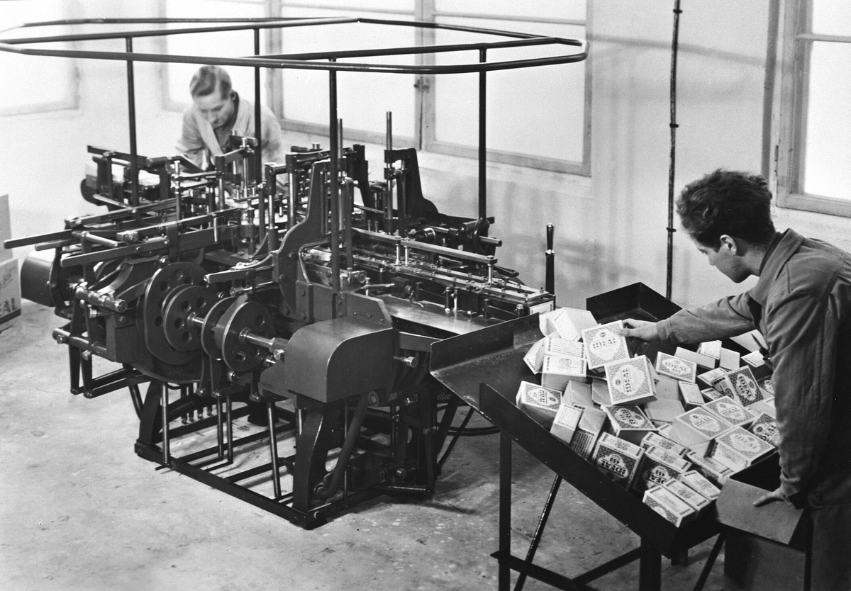 Anno Domkirkeodden, INTERIØR FABRIKK, 2 MENN VED PAKKEMASKIN, IDEAL FLATBRØDFABRIKK A/S i Hamar. Emil Nordby startet fabrikken i 1924 og klarte å fremstille tynt flatbrød maskinelt. Ideal lyktes med å eksportere til utlandet i en vanskelig tid, blant annet til de norske miljøene i USA. Maskinene lagde de internt.