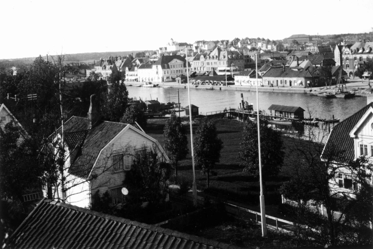 Hermann Christian Neupert, Fredrikstad, Østfold. 1930. Oversiktsbilde over byen. Villaer i forgrunnen med hager ned til elva. Fortøyde båter langs elva. Bybebyggelse i bakgrunnen.