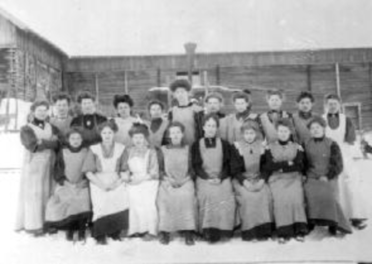 Anno Domkirkeodden, Gruppe elever fra husholdningskurs på Eskerud, Helgøya. Den gamle tømmerlåven i bakgrunnen. Damene er sittende fra venstre : Tora Kleven, Astrid Lier (Skallerud), Inger Larsen (Amundsen), Ella Staff, lærerinne frøken Deienvold, Lovise Maurud (Narmo), Olga Ekeberg Olsen, Pauline Larsen. Stående bak …