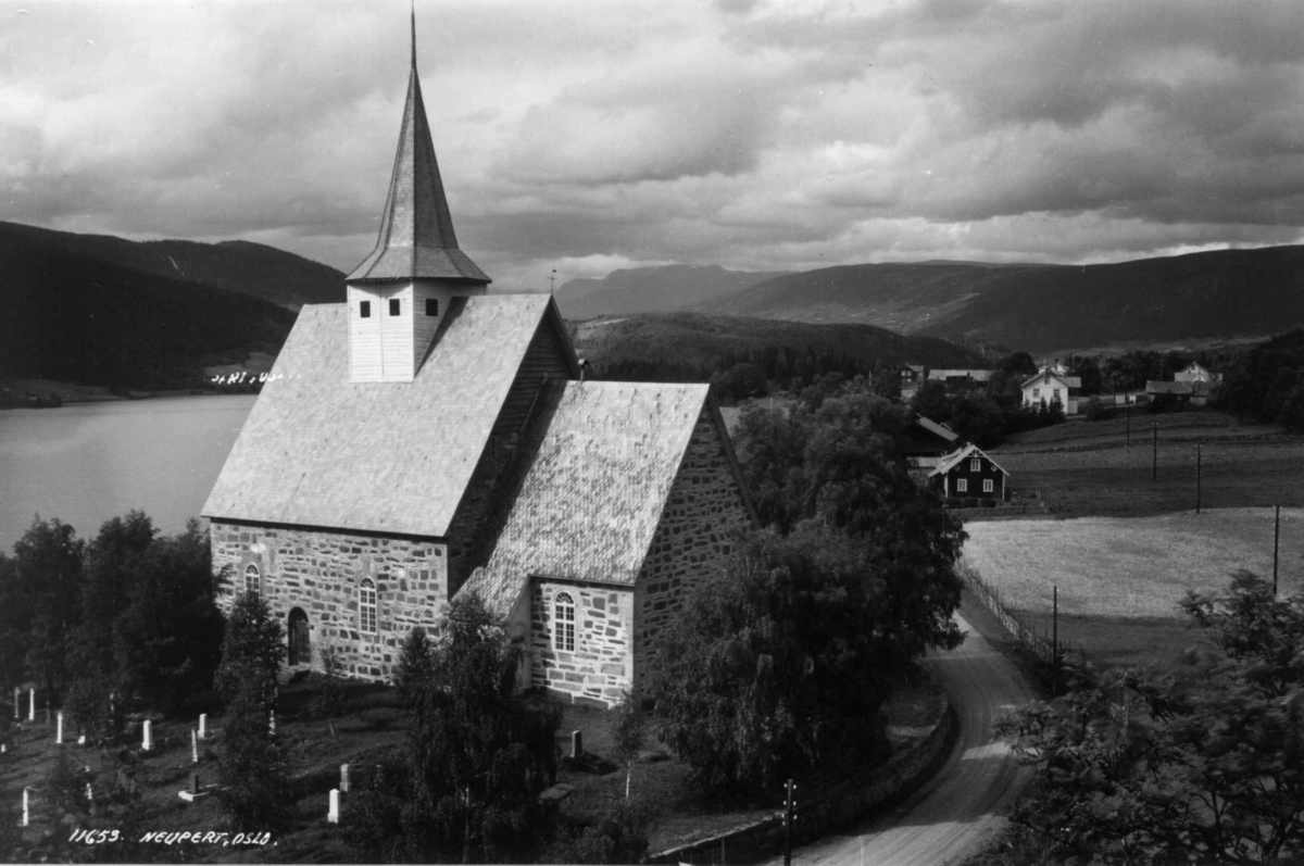 Hermann Christian Neupert, Vestre Slidre kirke, Oppland 1934. Steinkirke bygget 1150-1200. Omgitt av kirkegård med vei forbi. Utsikt mot Slidrefjorden og jordbruksbygd med gårdsanlegg. Åser og fjell i bakgrunnen.