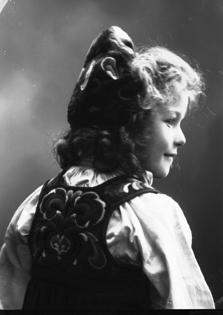 Solveig Lund, Studioportrett av pike i drakt og lue, fotografert bakfra.
