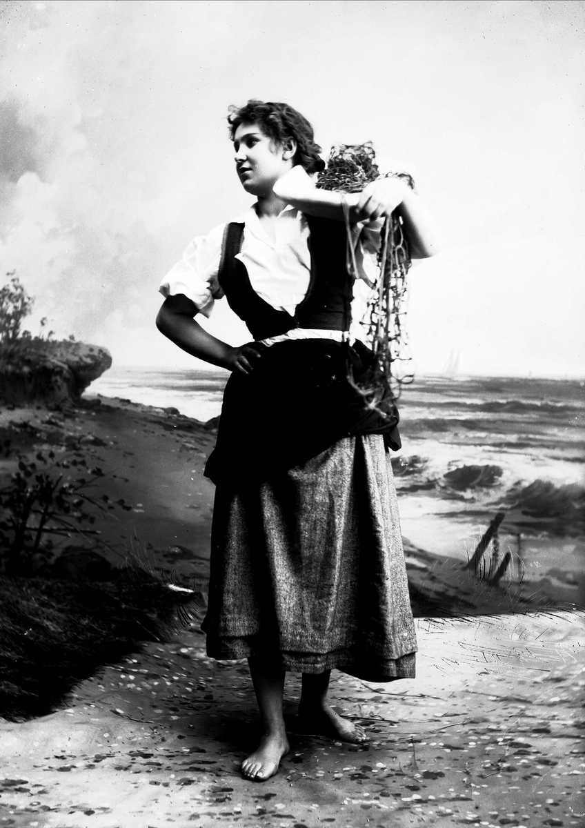 Solveig Lund, Studioportrett av kvinne i drakt som holder trau med fiskegarn på den ene skulderen. Hun ser ut mot venstre i bildet. Bakgrunnen er malt med sjø og sand.