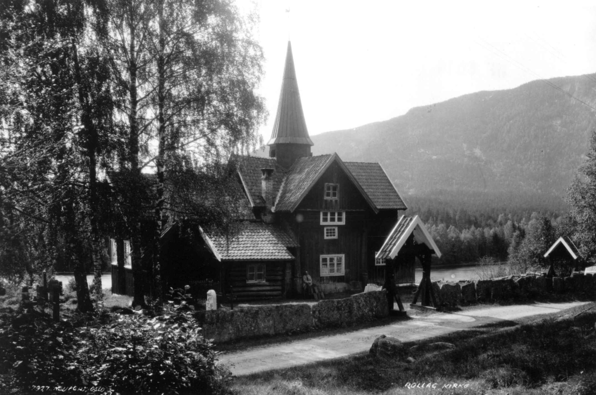 Hermann Christian Neupert, Rollag kirke - Rollag, Buskerud. 1930. Kirkegård. Trær. Gravsteiner. Mann sittende ved inngangen. Skog og åser i bakgrunnen.