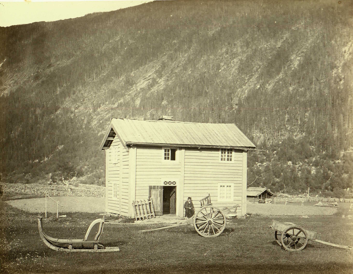 Henry Rosling, Gjestgivergården Dale i Vestfjordddalen i Tinn i Telemark, bygget i 1830-årene. Landskap nær Gaustatoppen. I forgrunnen trebygning, bolighus, ant. hos Ole Torgersen Dahl (se også NF.15003-027, ant. samme hus). Mann sitter utenfor huset, ulike hestekjøretøyer er plassert i forgrunnen. Fra serie nors…