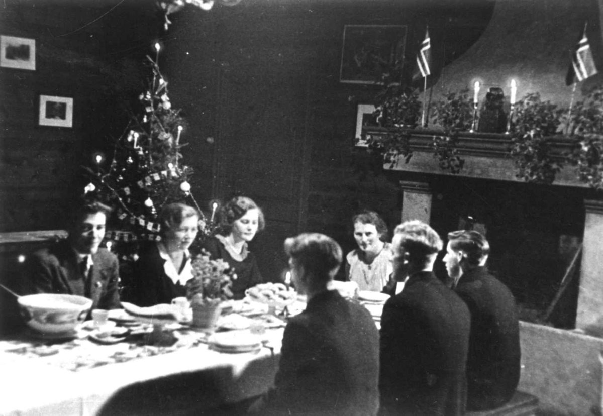Anna Grostøl, medlemmer av Anna Grostøls familie sitter rundt et dekket spisebord foran peisen. Et juletre i bakgrunnen. Vest-Agder 1941-42.