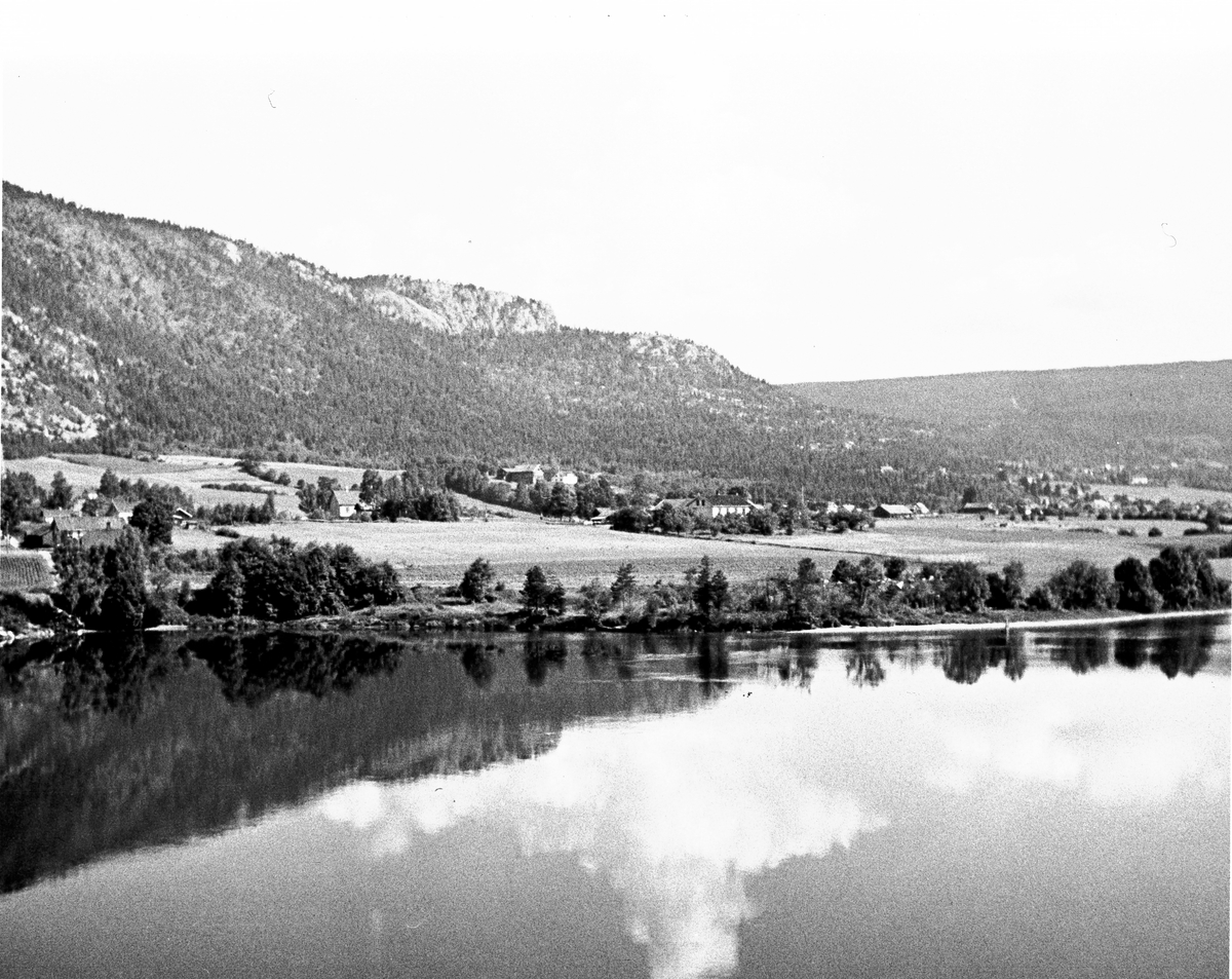Anders Beer Wilse, Gårdsbruk og åkre med fjellet i bakgrunnen, speiler seg i vannet langs landskapet. Antatt Mjøndalen. Fotografert 1940.