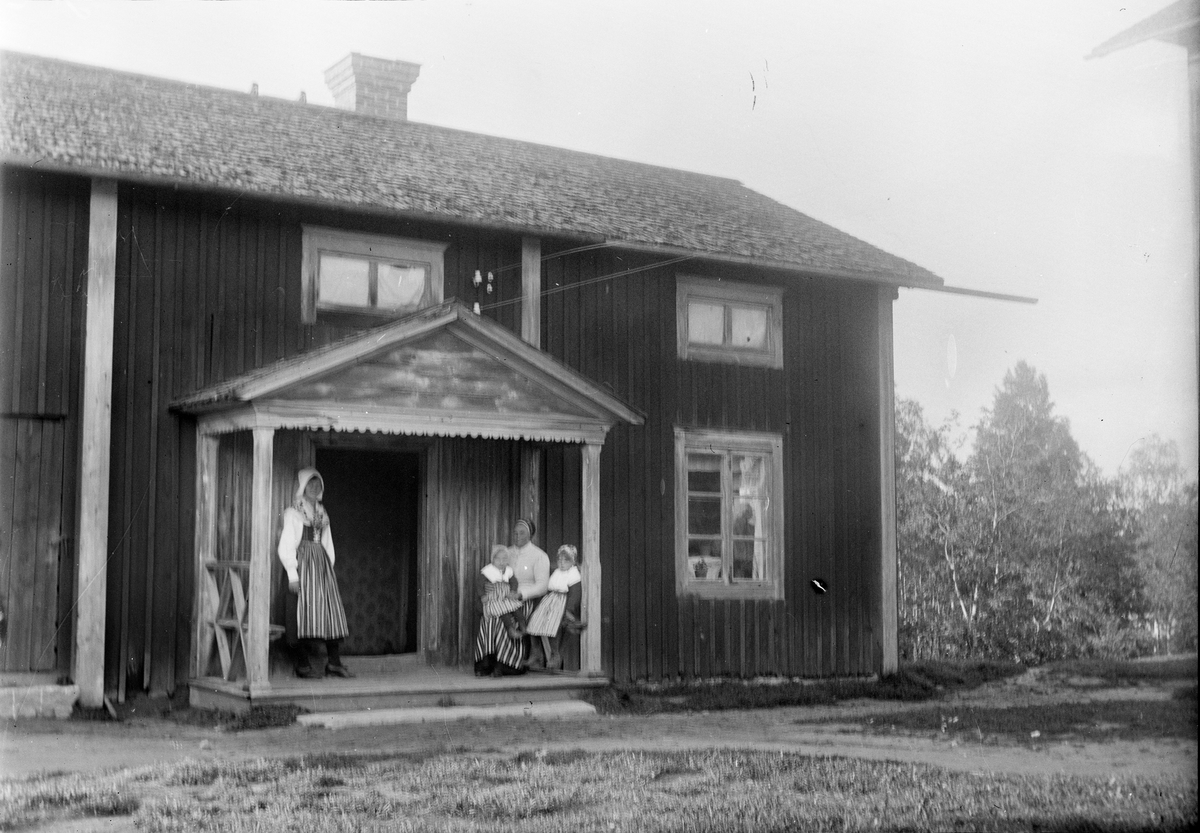 Okänd,, Two women and two little girls dressed up folk costume in front of residential buildings, probably in Dalarna