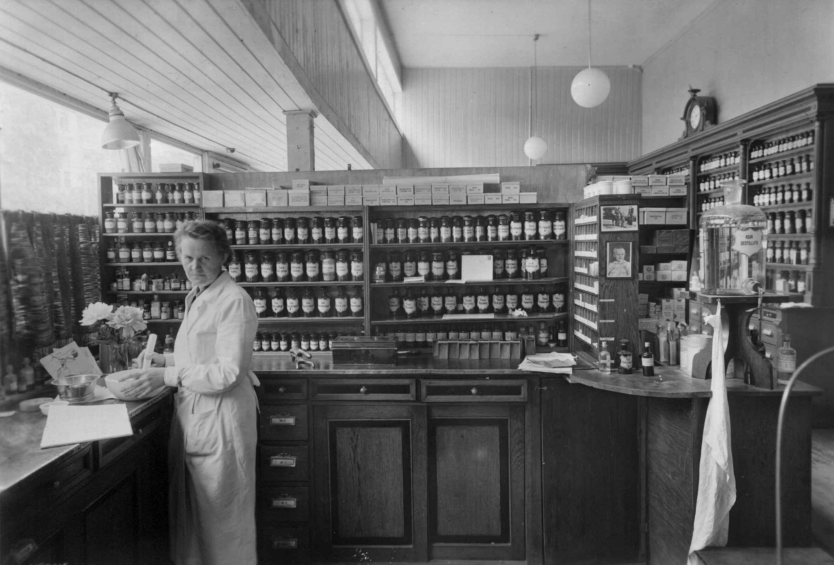 Hermann Christian Neupert, Rådhusapoteket, Oslo. 1935-1937. Interiør. Disk med skuffer. Kvinne ved disken. Hyller med aptokekervarer i flasker