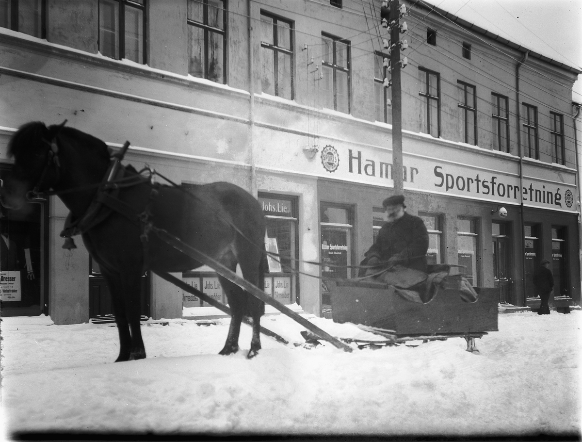 Ragnvald Andersen, Hesten Trygg med Oluf Danielsen i slede i Torggata, foran Hamar Sportsforretning og Johs Lie Skrædder. Hamar.