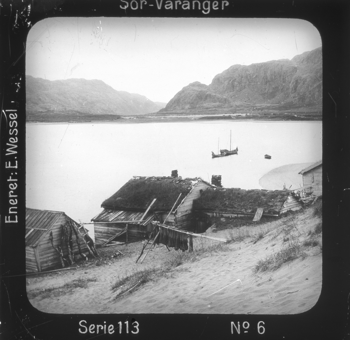 Ellisif Rannveig Wessel, Grense Jakobselv, Sør-Varanger, Finnmark, ved Jakobsviken. I forgrunnen gårdsbebyggelse, fiskegarn tørkes. Motivet har nr.6 i lysbildeforedraget kalt "I lappernes land - Sør-Varanger", utgitt i Nerliens Lysbilledserier, serie nr 113.