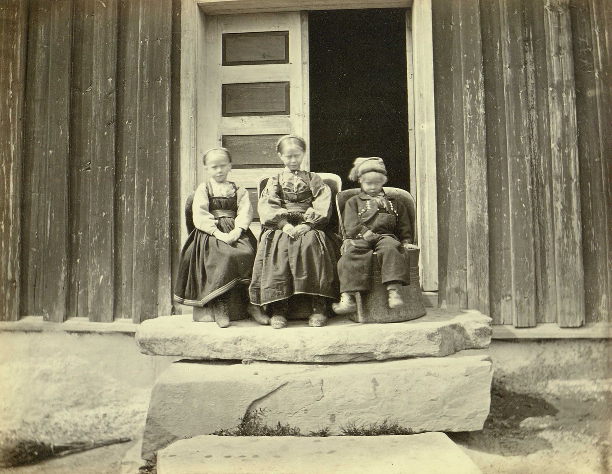 Henry Rosling, Barnegruppe i folkedrakter, Heddal, Notodden, Telemark. To piker og en gutt sitter på kubbestoler foran inngangsdør. Fra serie norske landskapsfotografier tatt av den engelske fotografen Henry Rosling (1828-1911).