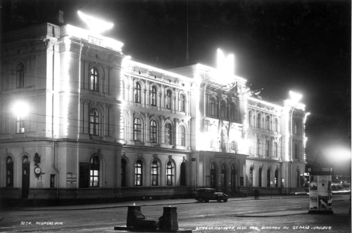 Hermann Christian Neupert, Håkon VII's 25. års jubileum. 1930. Østbanen i Oslo opplyst. Gate. En bil foran stasjonsbygningen..