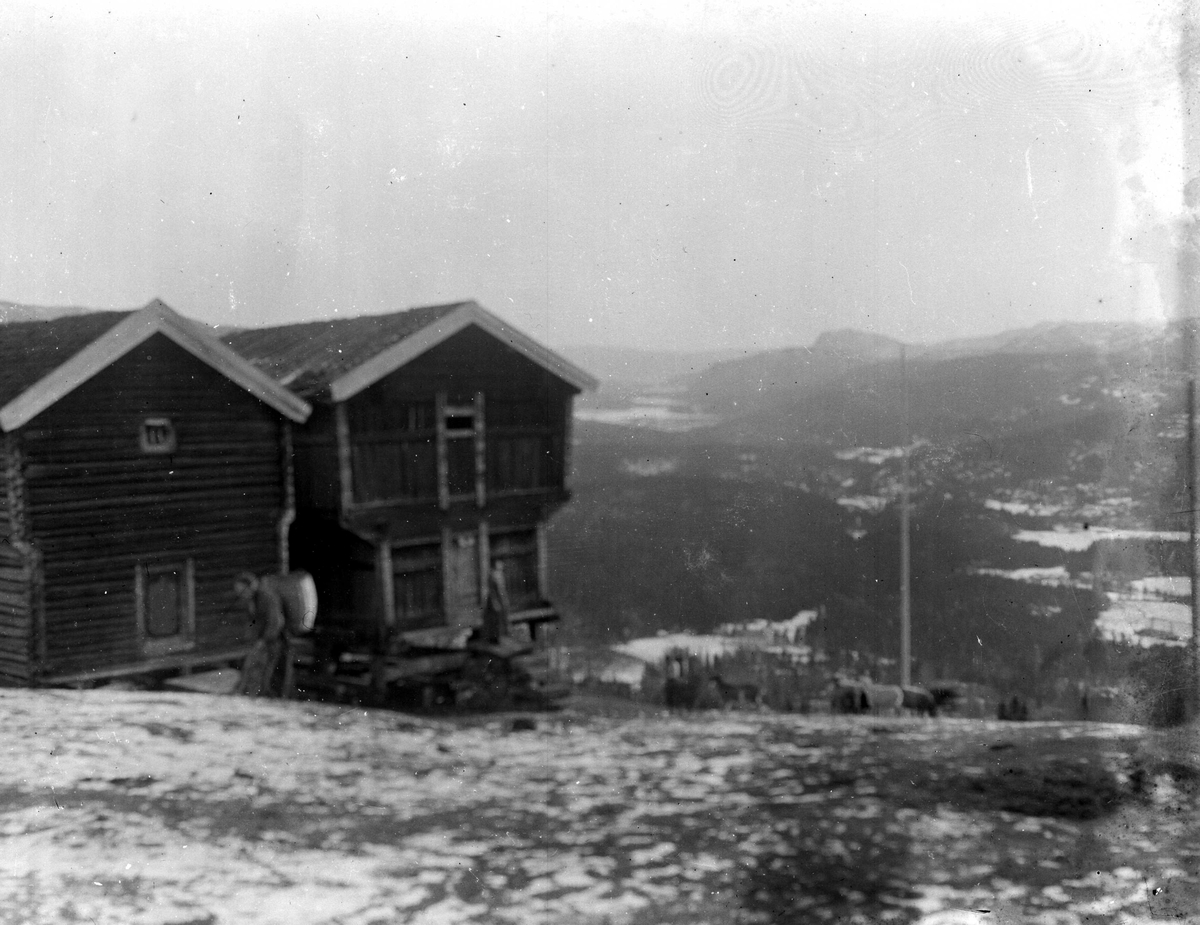 Juliane Fredrikke Langberg, Fra Eggedal, Sigdal, på besøk hos Th. Kittelsen. Høytliggende gårdstun med uthus, loft/stabbur. Kvinne i lokal folkedrakt bærende på en stor kurv (kont?), ant. med høy, foran uthusene. Samme motiv, skarpere, se NF.15386-100. Fra malerinne Juliane Fredrikke Langbergs samling, tatt 1895-1910 med moti…