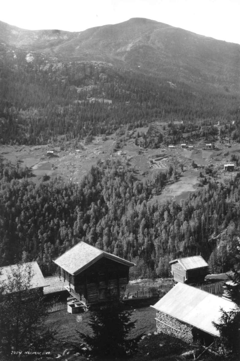 Hermann Christian Neupert, Rollag - Buskerud? 1930. Oversiktsbilde. Gårdstun med loft i forgrunnen. Landskap med gårder, skog og fjell i bakgrunnen.