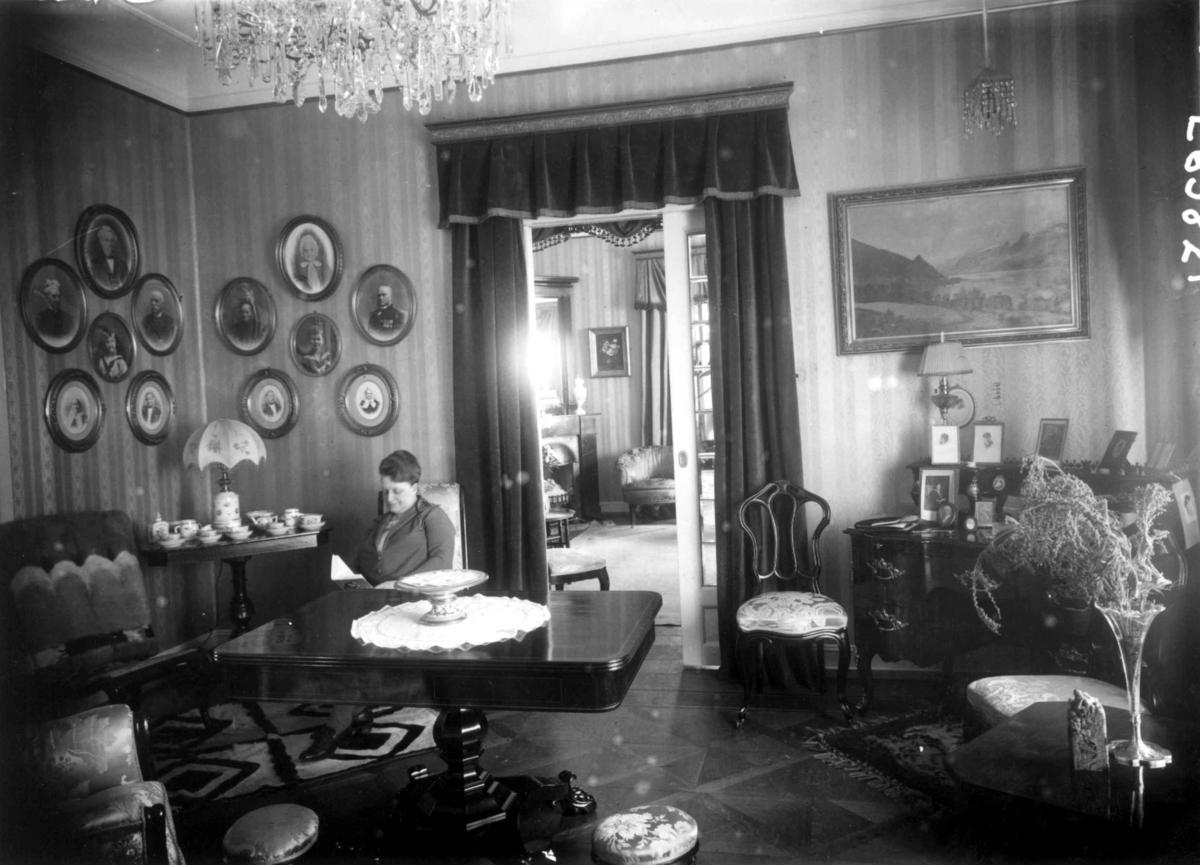 Hermann Christian Neupert, Trapp-Meyer. Stueinteriør 1925. Stoler og bord. Kvinne med bok. Portretter og maleri på veggene. Lysekrone.