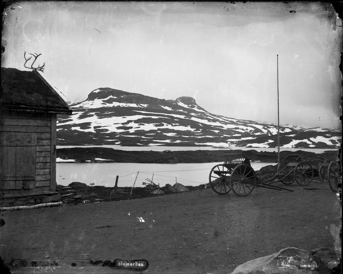 Marthinius Skøien, Hestekjøretøyer og bygning ved innsjø med fjell i bakgrunnen, Telemark.