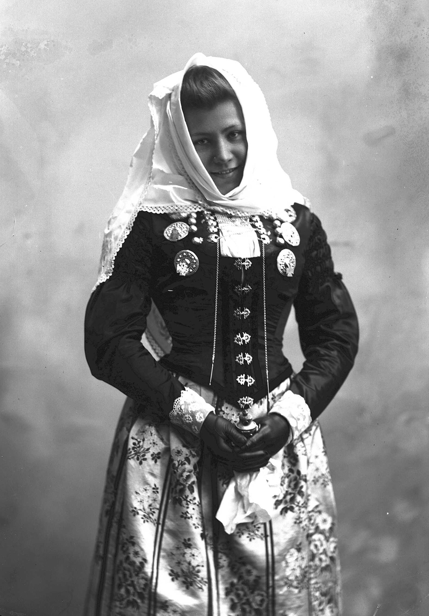 Solveig Lund, 3/4 dels studioportrett av kvinne i drakt og skaut. I hendene holder hun en lukteflaske. Hun er avfotografert mot en nøytral bakgrunn.
