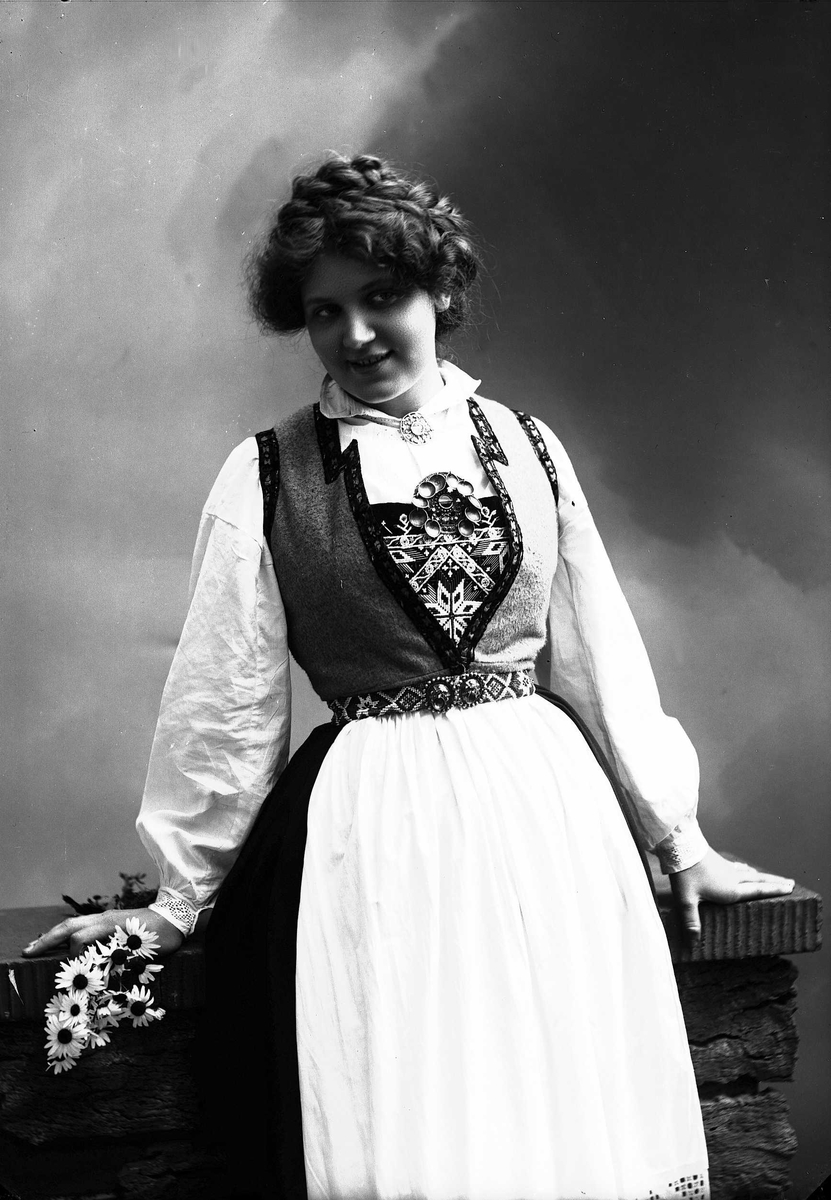 Solveig Lund, 3/4 dels studioportrett av kvinne i drakt og oppsatt hår, som lener seg mot et gjerde. I høyre hånd holder hun en bukett prestekrager. Bakgrunnen er malt med himmel og skyer.