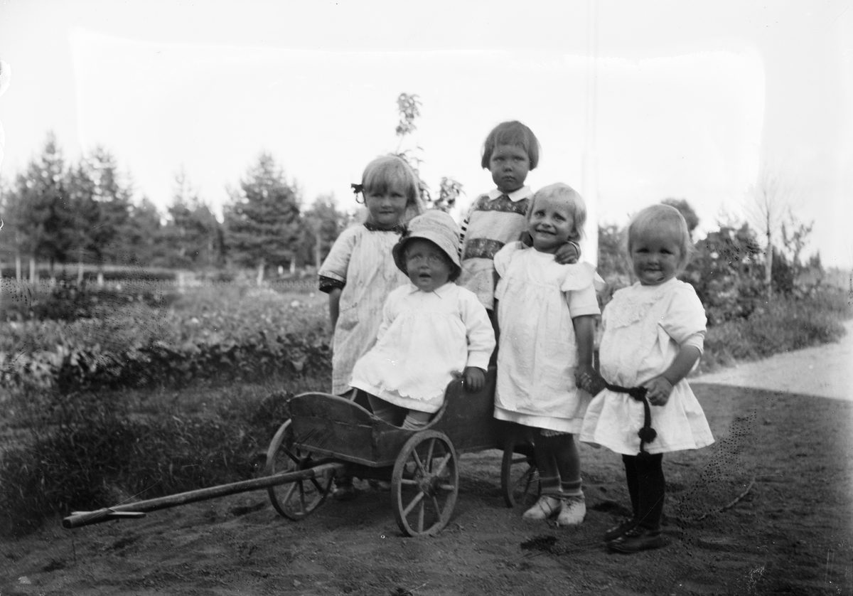 Okänd,, Marit Liljefors med fyra andra små flickor i trädgård, en sitter i en kärra. Sannolikt i Sverige