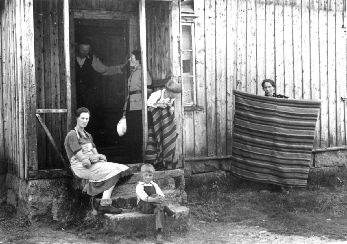 Anna Grostøl, Anstein Åvitsland med flere samlet utenfor inngangspartiet til huset hans. Datteren viser frem et gammelt sengeteppe. Fjotland, Kvinesdal, 1941.