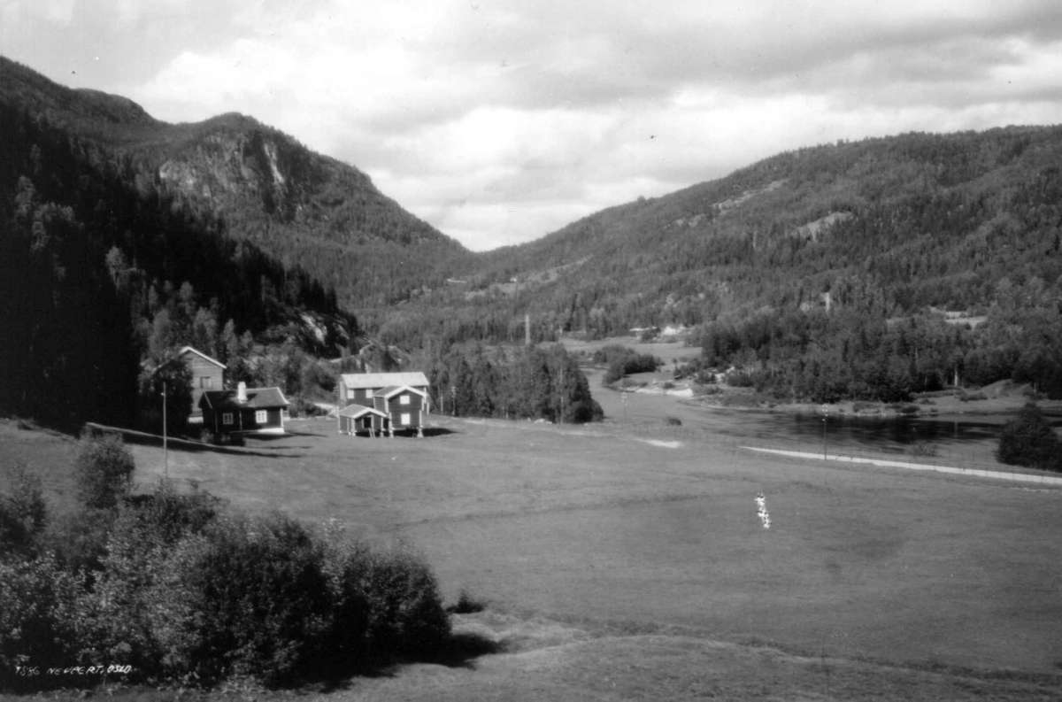 Hermann Christian Neupert, Landskap, Buskerud. 1930. Oversiktsbilde med gårder, vei, elv og skog. Åser i bakgrunnen.