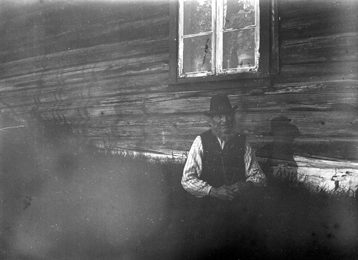 Juliane Fredrikke Langberg, Portrett av bonde i Eggedal, eldre mann med vest og skjorte, hatt og skjegg sittende foran umalt husvegg med vindu. Fra malerinne Juliane Fredrikke Langbergs (1856-1930) samling, tatt ca.1895-1910 med motivkrets slekt og venner samt steder tilknyttet samvær med disse.