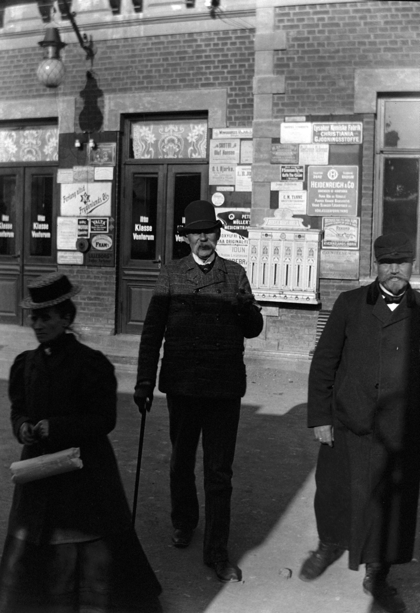 Ole Hansen Løken, 3 UKJENTE, MANN, HATT OG STOKK, REKLAMEVEGG, CIGAR OG CIGARETT AUTOMAT. Jernbanestasjon.