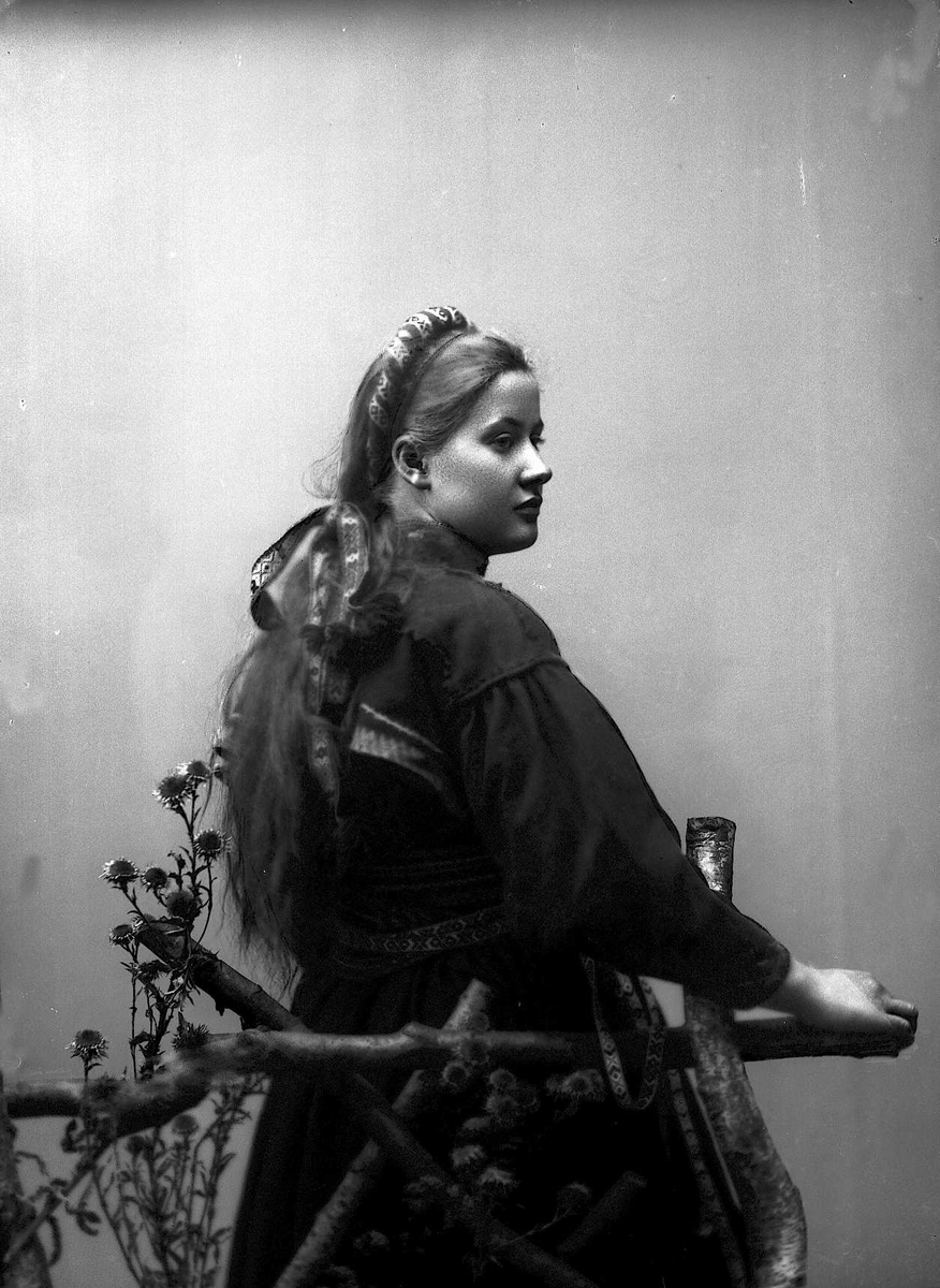 Solveig Lund, 3/4 dels studioportrett av kvinne i drakt og band i håret. Hun sitter med ryggen til fotografen bak et gjerde av trestokker.