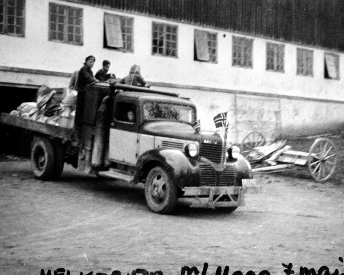 Anno Domkirkeodden, Ivar Vangens lastebil med melkespann som kjørte melkeruta på Helgøya. Dodge modell 1939 er utstyrt med flagg. Sjåfør er Bjarne Strøm. Utenfor låven på Hovelsrud, Helgøya i fredsdagene i mai 1945.