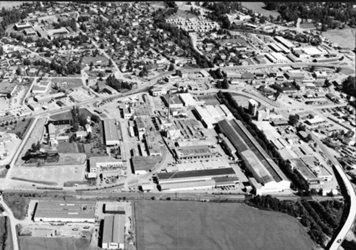 Widerøes Flyveselskap A/S, Flyfoto over Berger Langmoen A/S, Norske Skog, Brumunddal. Fra venstre er etterbehandlingsavdeling for wallboardfabrikken, øverst sag, fyrhus, flissilo, etterbehandling trelast, rekvisitaavdeling og nærmest jernbanen er høvleriet. langs elva er virkesmagasin med tretørke øverst, parkettfabrikken på…