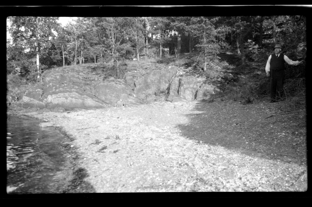 Rolf Sr. Sundt, Ukjent mann ved strand. Hus på naturtomt i bakgrunnen.