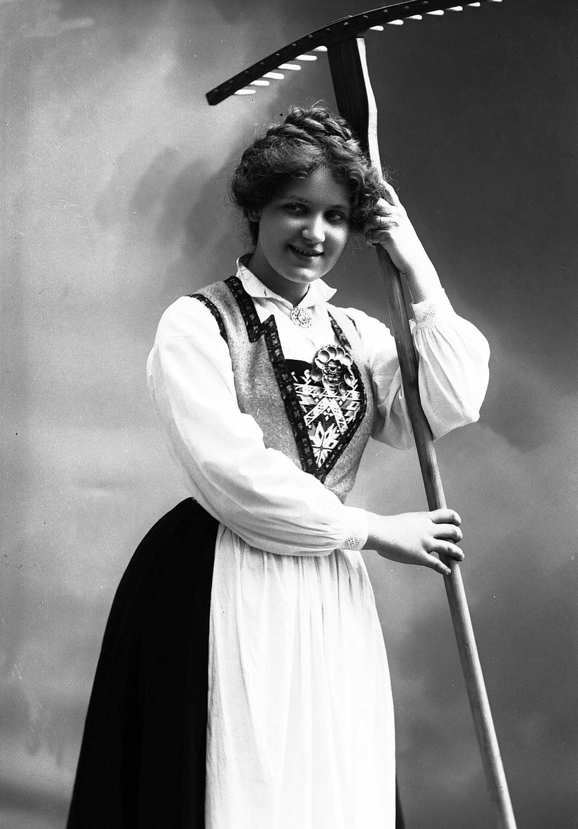 Solveig Lund, 3/4 dels studioportrett av kvinne i drakt, med oppsatt hår. Hun lener seg mot en rive. Bakgrunnen er nøytral.