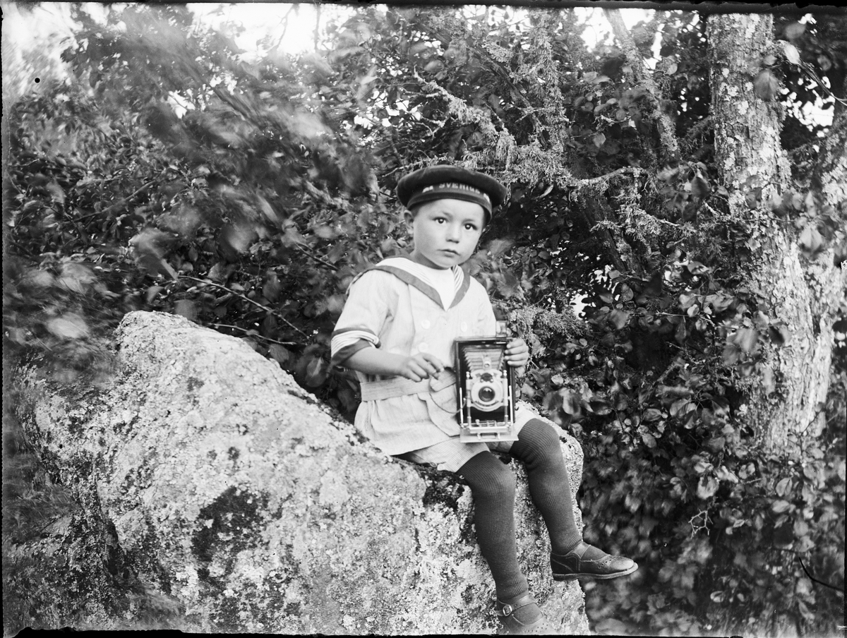 Skötsner-Edhlund, Little boy dressed in sailor costume sits on a stone with a camera in his lap, Östhammar, Uppland