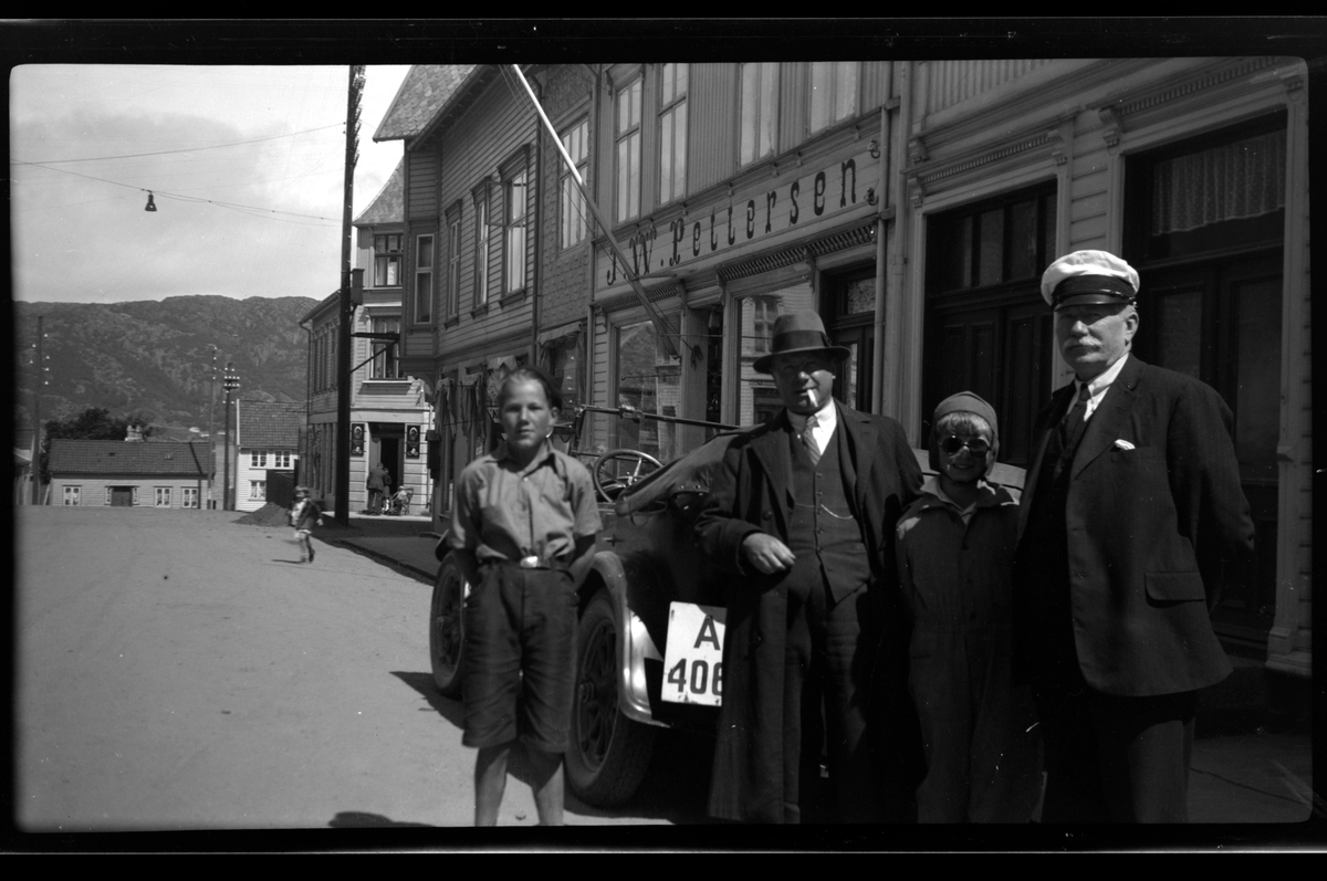 Rolf Sr. Sundt, I Storgaten i Farsund står Bjørn Reymert, Finn og Rolf jr. Sundt og Christen Reymert foran en bil, som står parkert. Bebyggelse med butikker og kiosker i bakrunnen.