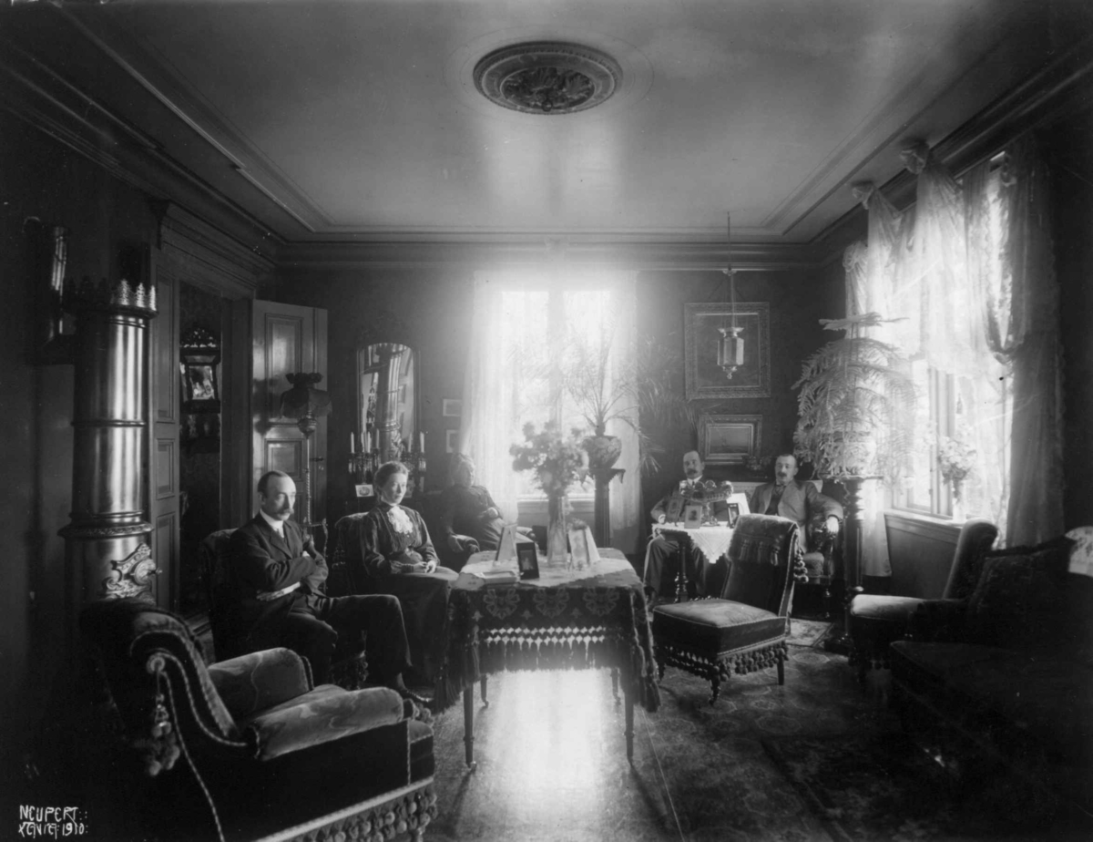 Hermann Christian Neupert, Holm. Stueinteriør. 1908-1910. To menn i en sofa. En mann og to kvinner sittende rundt et bord. Stueplanter og blomster.