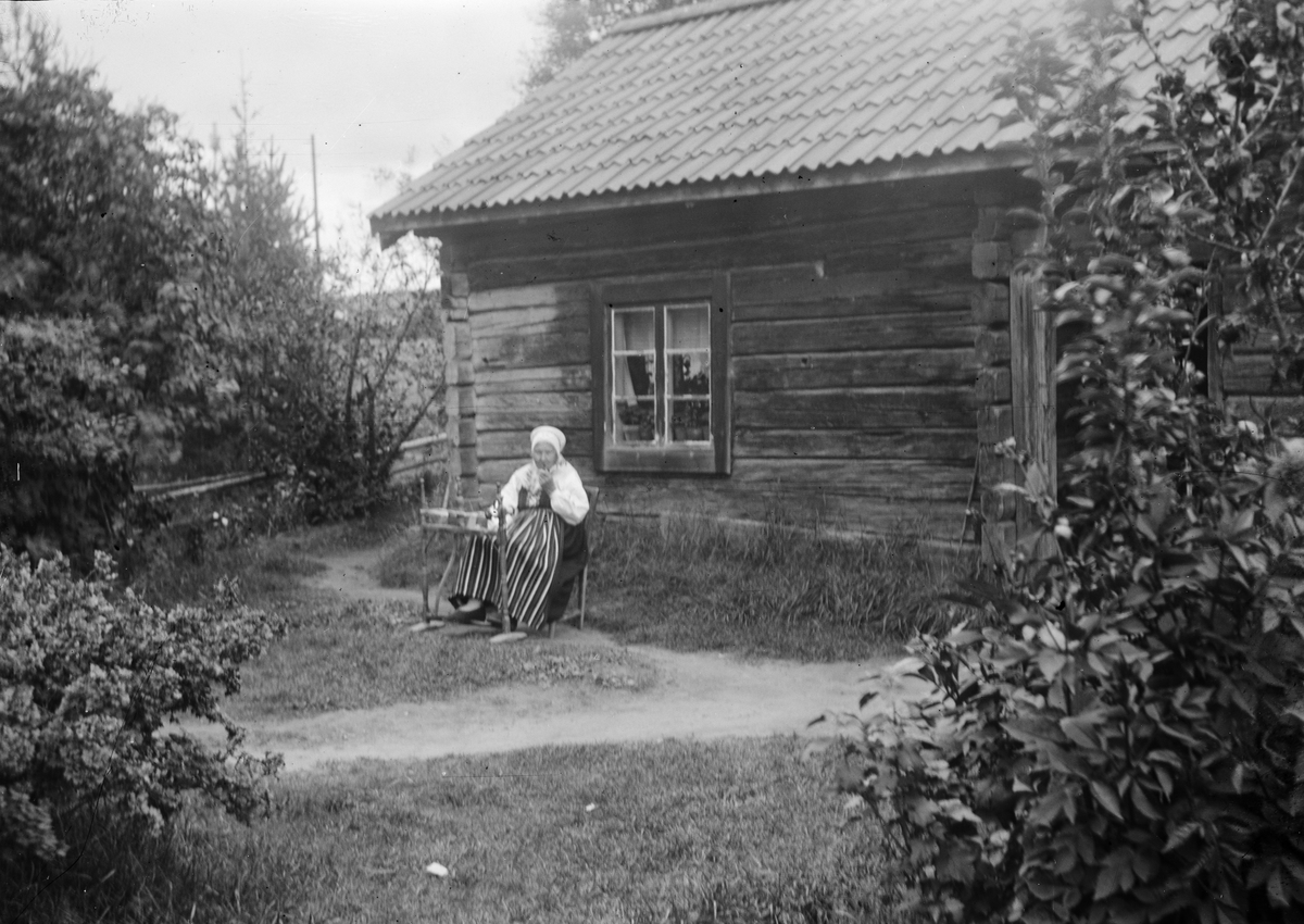 Okänd,, Elderly woman sitting outside a cottage dressed in folk costume and handworking, probably Dalarna