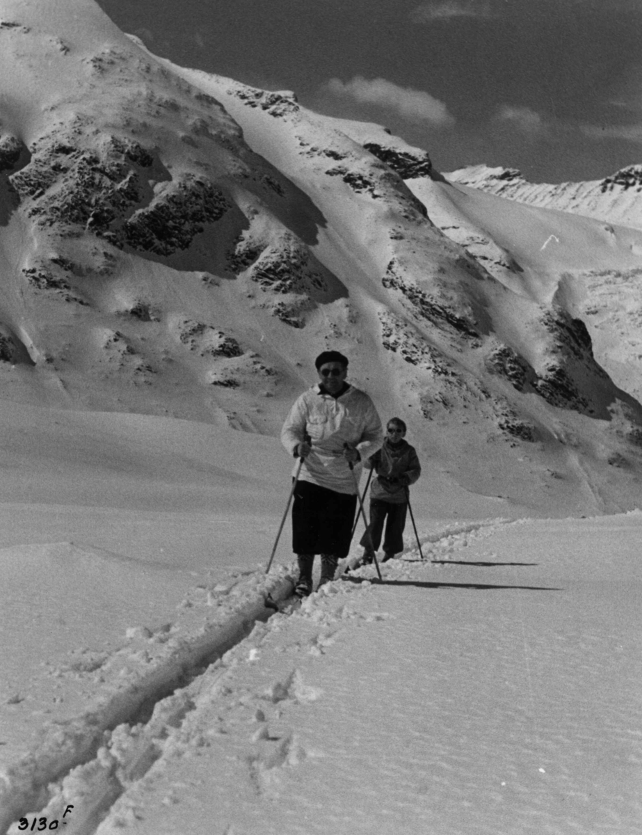 Anders Beer Wilse, Skiløpere ved Galdhøpiggen i Jotunheimen med snødekte fjell og sol.