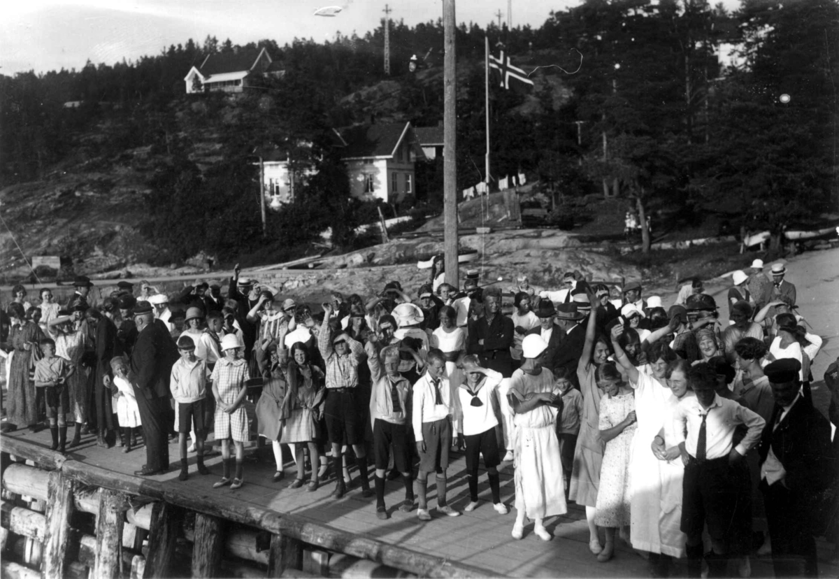 Hermann Christian Neupert, Hvitsten - Vestby, Akershus 1924. Oversiktsbilde. Festkledde mennesker samlet på brygge. Hus i bakgrunnen og flagg.