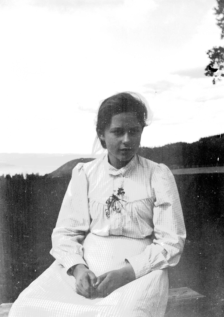 Juliane Fredrikke Langberg, Portrett av jente, Emily Holm, fotografens niese, sittende på en en benk i åpent terreng, kledd i lys sommerkjole. Muligens tatt i Hurum eller Rakkestad, der faren var prest. Fra malerinne Juliane Fredrikke Langbergs (1856-1930) samling, tatt ca.1895-1910 med motivkrets slekt og venner samt steder …