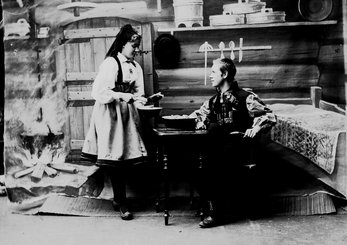 Solveig Lund, Studioportrett av mann og kvinne i nasjonaldrakter. Kvinne holder sleiv med mat i hånden og serverer mannen som sitter ved et bord. Bakgrunnen er malt og viser seterinteriør. Til venstre står kjelen på ildstedet og til høyre står seng og vegghylle med tiner.
