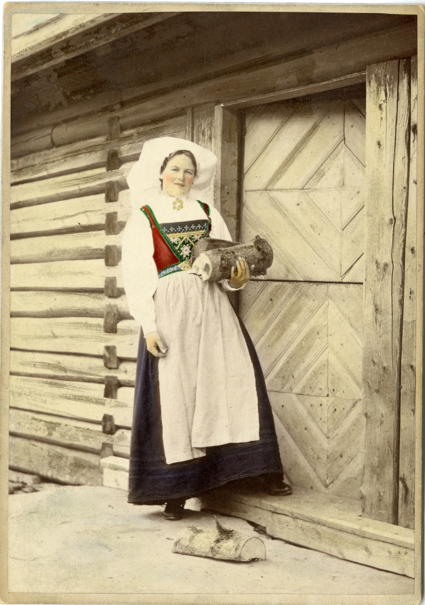Solveig Lund, Kolorert fotografi av kvinne med drakt, bærer ved står foran dør. Hardanger.