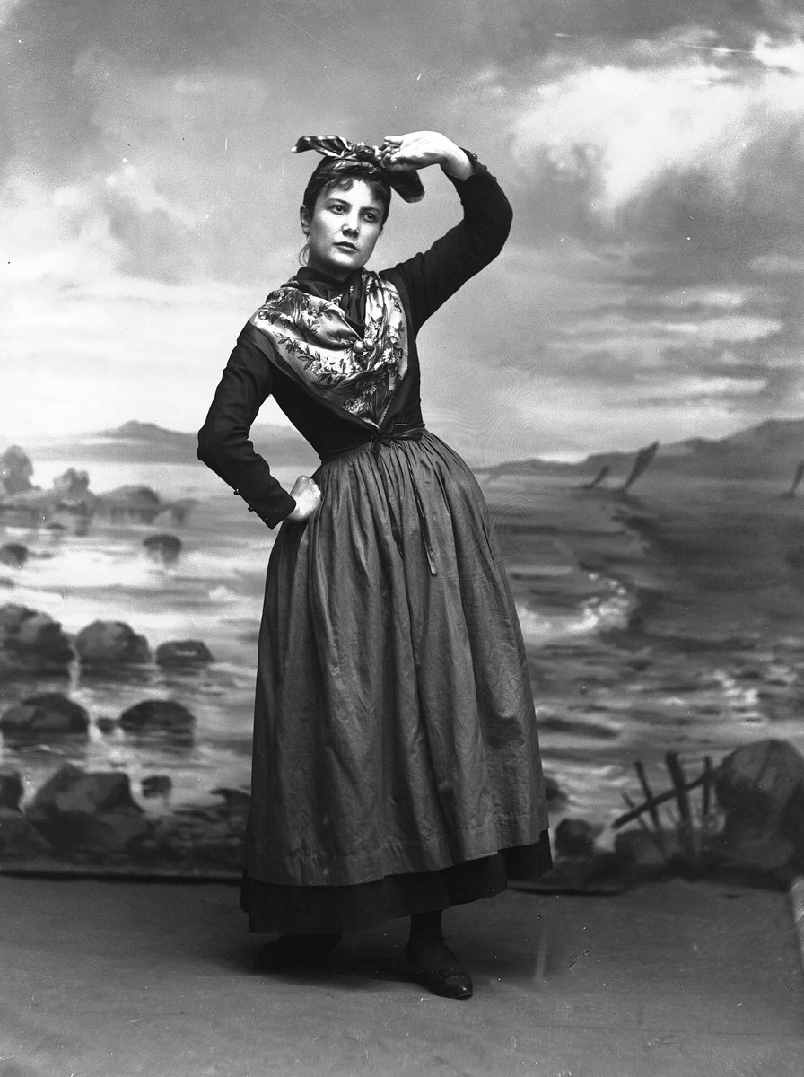 Solveig Lund, Studioportrett av kvinne i drakt og tørkle på hodet som speider. Bakgrunnen er malt med sjø, himmel og steiner.