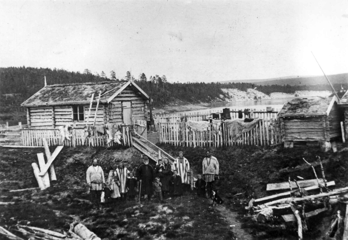 Konrad Nielsen, Seter i Assebakte. Inngjerdet tømmerhus, et stabbur utenfor. Nedenfor gjerdet med skinnfeller, står barn og voksne.