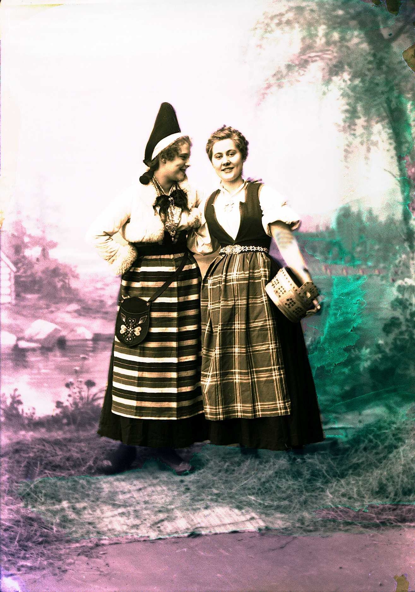 Solveig Lund, To kvinner i drakt. En holder en ambar under armen. Malt bakgrunn med landskap og seter, høy på gulvet foran teppet.