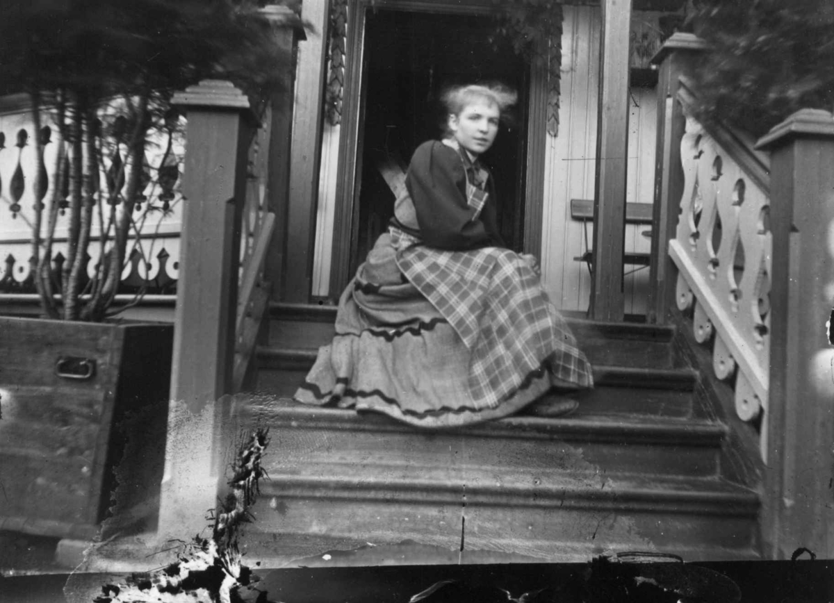 Hermann Christian Neupert, Drakt 1908-10. Kvinne sittende i en trapp. Veranda i balgrunnen.
