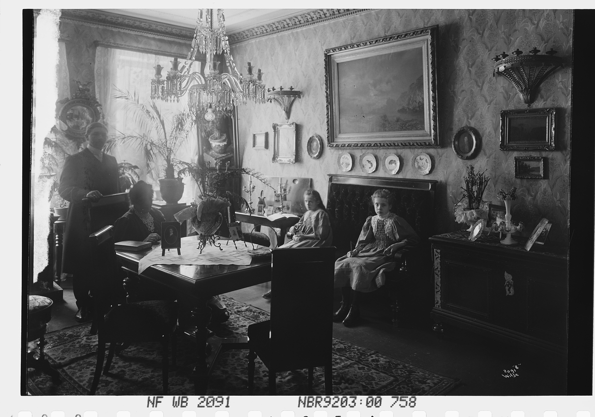 Anders Beer Wilse, Fru Simonsens stue i all sin prakt. To piker, antatt døtre, sitter i sofaen, fru Simonsen ved bordet og en vel antrukket yngre herre står bak fruens stol.