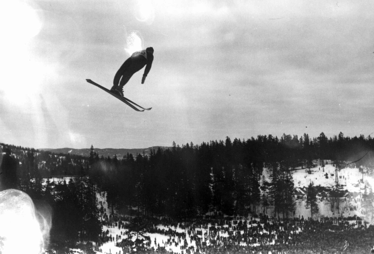 Hermann Christian Neupert, Ski. Hopprenn. Holmenkollen, Oslo 1934. En hopper i svevet. Publikum ses på sletta. Nordmarka i bakgrunnen