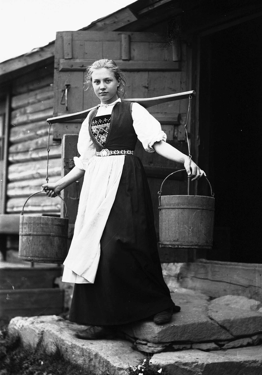 Solveig Lund, Pike i Hardangerdrakt står foran dør til seter. Bærer på to vannbøtter.
